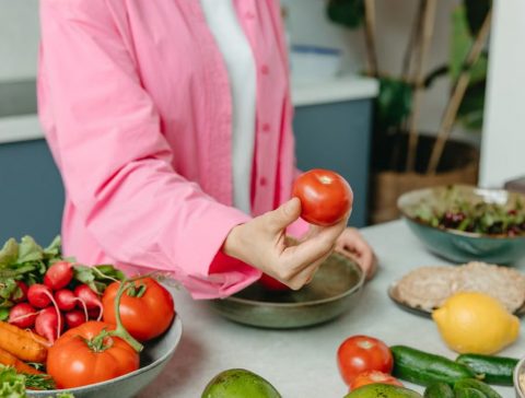 Person in pink overshirt holding a fresh tomato with various organic vegetables on a countertop.